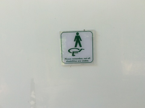 Sign saying "Please remember, not all disabilities are visible" showing a stick figure standing upright and a wheelchair logo which is upside down. I have no idea why the wheelchair user is upside down!