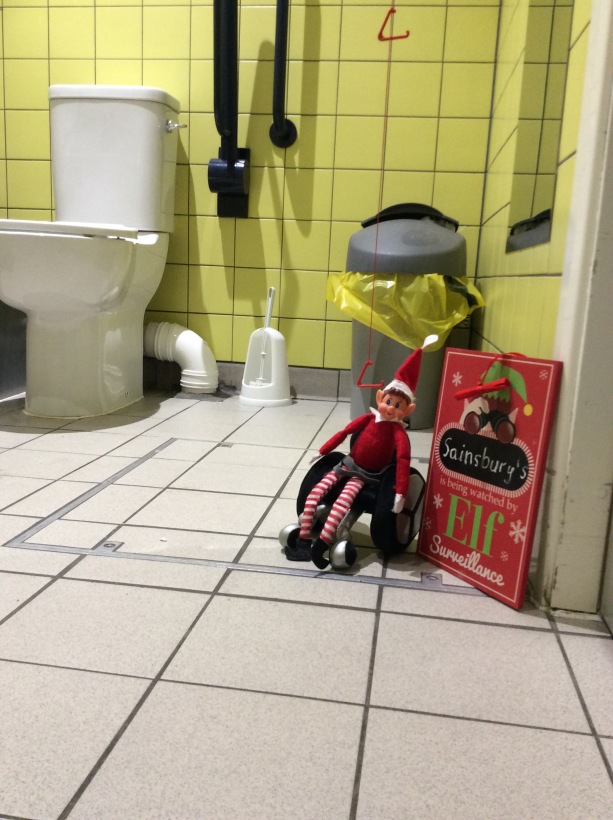 Elf in wheelchair next to a sign saying "Sainsburys is being watched by Elf Surveillance".