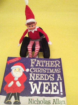 Alfie the Elf, sat in his wheelchair, with the book "Father Christmas Needs A Wee" in front of him.