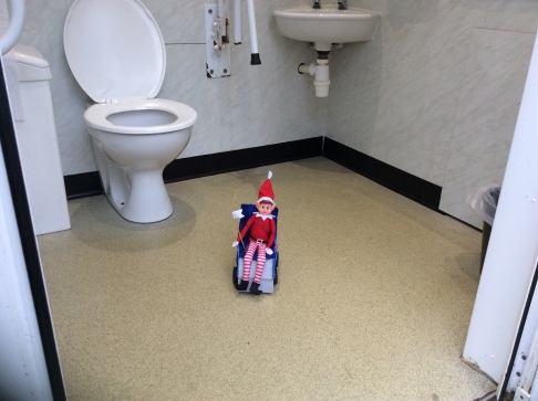 Toy Elf in a toy powered wheelchair, on the floor of a disabled toilet.