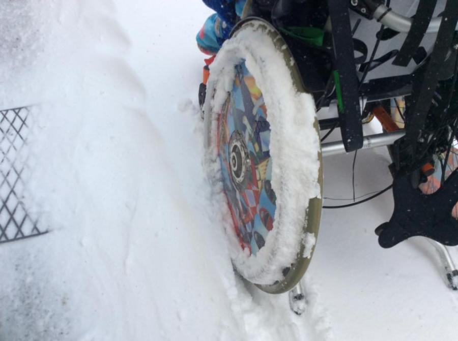 Rear view of a wheelchair in the snow, with the focus being the wheel of the chair absolutely covered with snow to a point where the push rim is no longer visible.