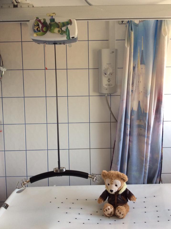 Photo showing a ceiling hoist and changing table. There is a bear wearing a jacket on the bench.