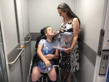 Wheelchair using child and his mum in a lift after shopping for toys. The boy is carrying two toys from the "Miles from Tomorrowland" show. Mum is carrying a Power Rangers T-Rex Zord. They are both smiling widely and looking at each other lovingly.