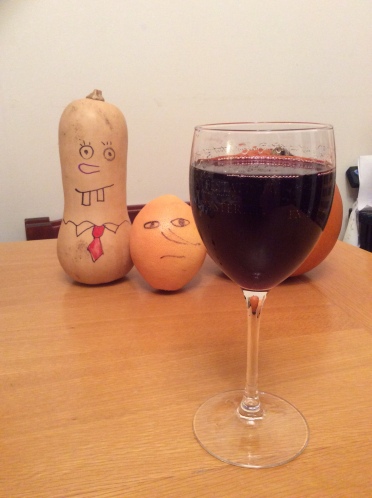 Glass of red wine in the foreground. Behind it is a butternut squash and a grapefruit, both of which have faces drawn on them. The butternut squash has two big gappy rectangular teeth, a big nose, round eyes, and a shirt collar with a red tie. He resembles Spongebob Squarepants. The grapefrui looks sternm with oval eys, a serious mouth and a big pointy nose. 