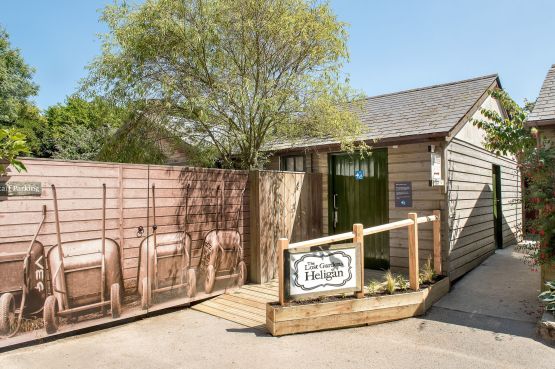 Photo shows a large wooden building with ramped access and a Changing Places logo on the door. The logo shows a wheelchair user, care with a changing table in frot f them and a ceiling hoist. There is a mural on the fence outside, the sky is bright blue and there are green trees in the background.