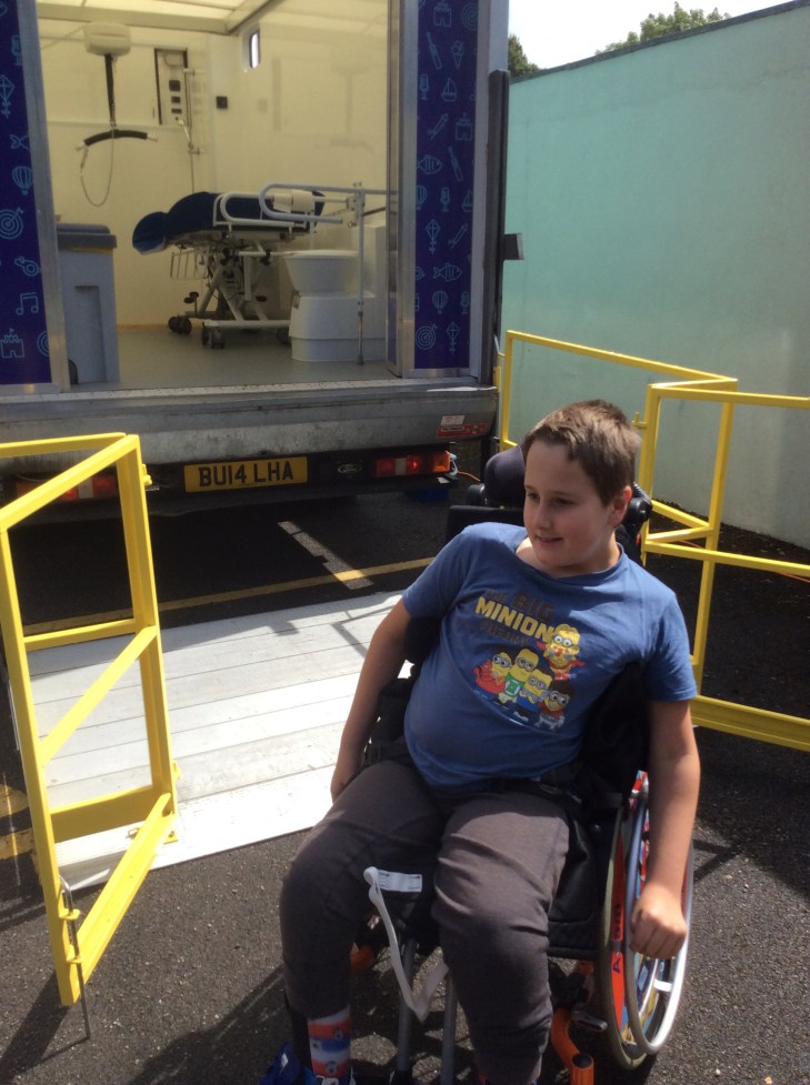 a smiling boy, wearing a blue t-shirt with Minions dressed as The Big Bang Theory characters, sat on his orange powered wheelchair. Behind him is a van with the tail lift down. Inside the van a toilet with rails, an adult sized changing table and ceiling hoist are all visible.