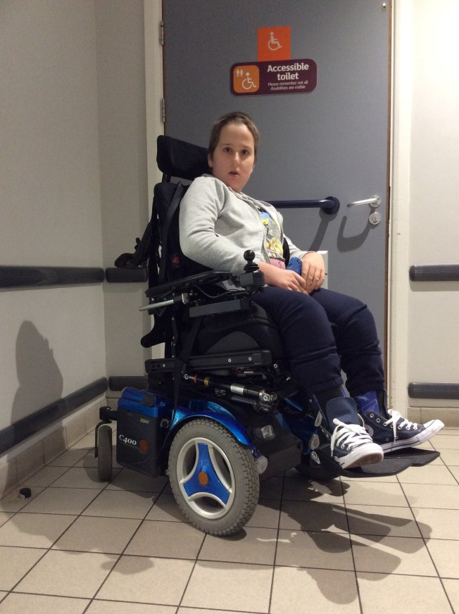 A 12 year old boy using a blue powered wheelchair is sitting outside of a supermarket's disabled toilet.The orange signage identifies it as Sainsbury's. There is a sign on the door saying 