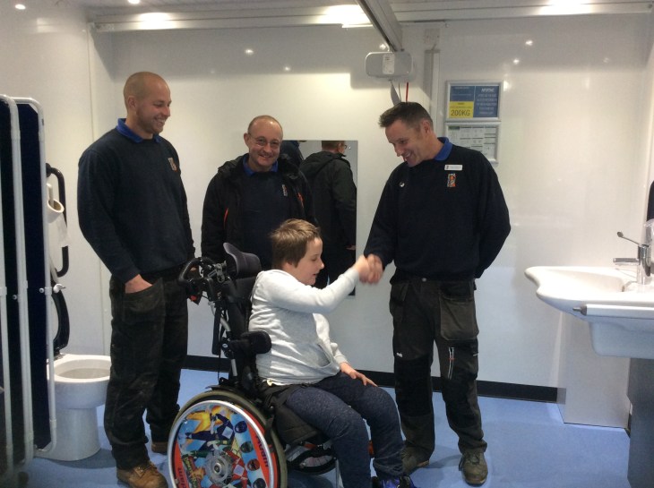 Three men wearing Newquay Zoo uniforms are standing inside the Changing Places toilets. A boy is shaking hands with one of them. The boy is wearing a light grey hooded top and dark grey trousers. He is using a manual wheelchair with Power Rangers on the wheels. All of them look very happy.