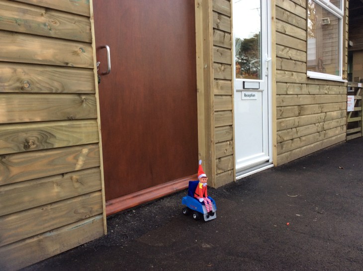 Toy Elf, sat in his powered wheelchair, outside a big wooden door, next to an office with 