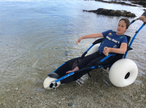 A boy using a Hippocampe beach wheelchair with sand wheels is in the sea. There are little black spots all over the sea. These spots are actually raindrops hitting the sea.