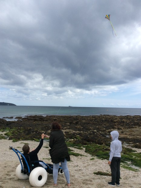 A boy using a sand wheelcair is flying a kite, aided by a lady and watched by another boy. The skies are grey, they are wearing warm clothes and it is clearly breezy from the height of the kite.